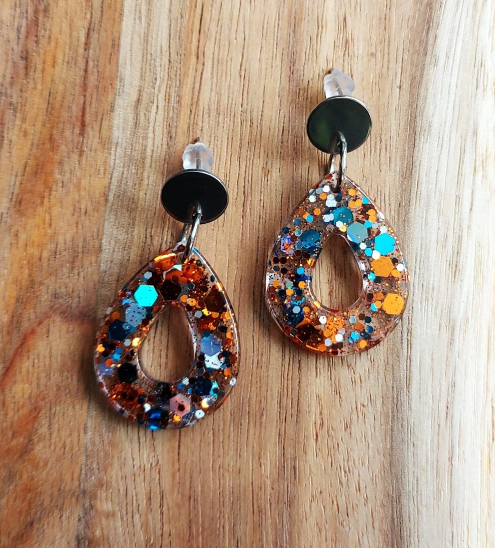 Small teardrop earrings, made in clear resin with a turquoise and copper chunky glitter mix added. They are fixed onto small flat silver tone stud tops, and displayed on a wooden board.