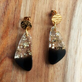 Geometric drop earrings made in resin with clear and gold flakes at the top graduating to smoky black at the bottom. They are fixed onto textured gold tone stud tops and displayed on a wooden board.