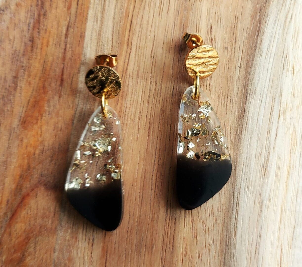 Geometric drop earrings made in resin with clear and gold flakes at the top graduating to smoky black at the bottom. They are fixed onto textured gold tone stud tops and displayed on a wooden board.