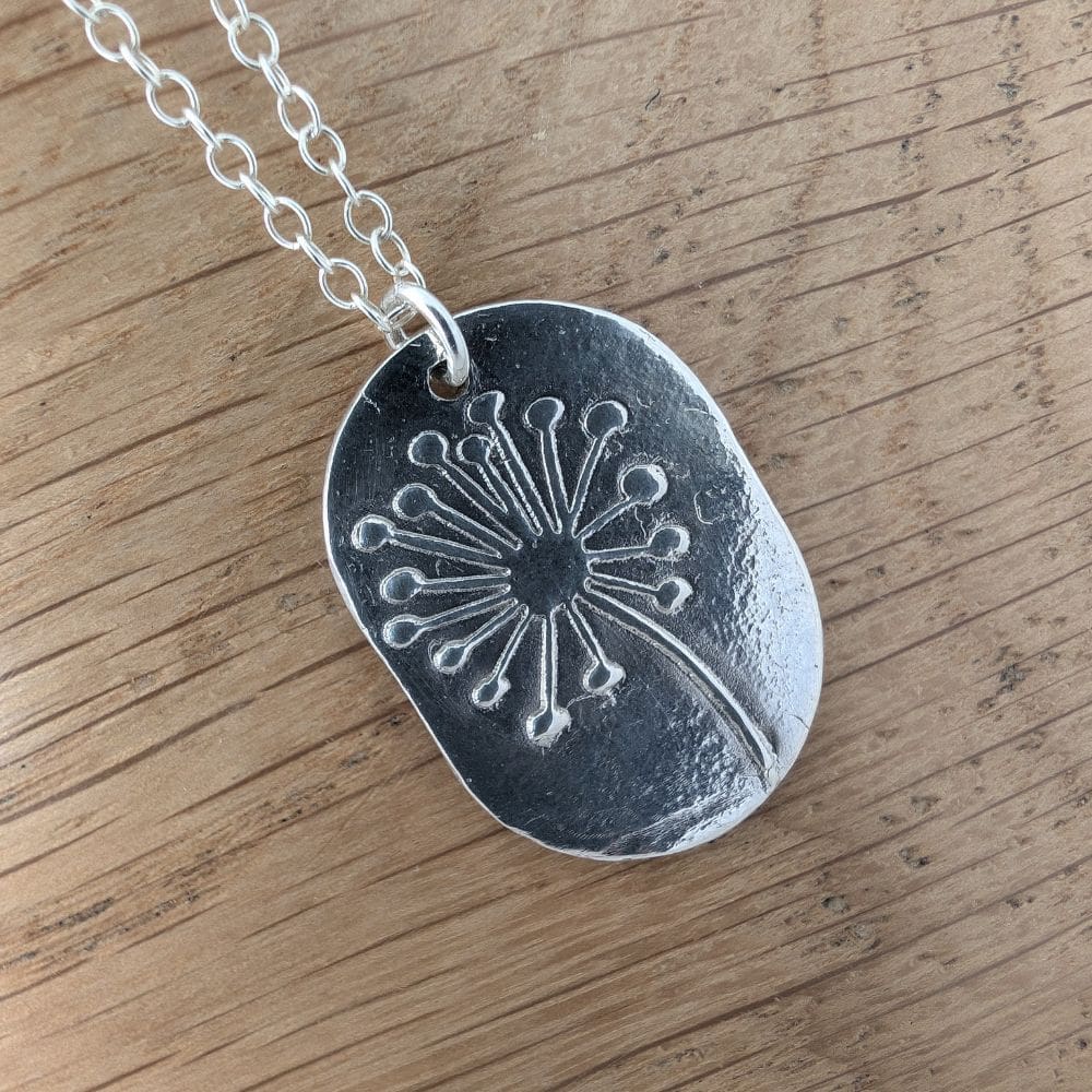 A dandelion clock imprint on a fine silver dog tag style pendant. Laid on a wodden background