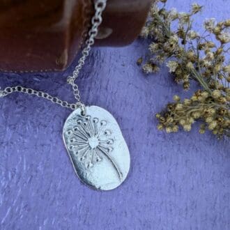 A dandelion clock imprint on a fine silver dog tag style pendant. Laid on a wodden background