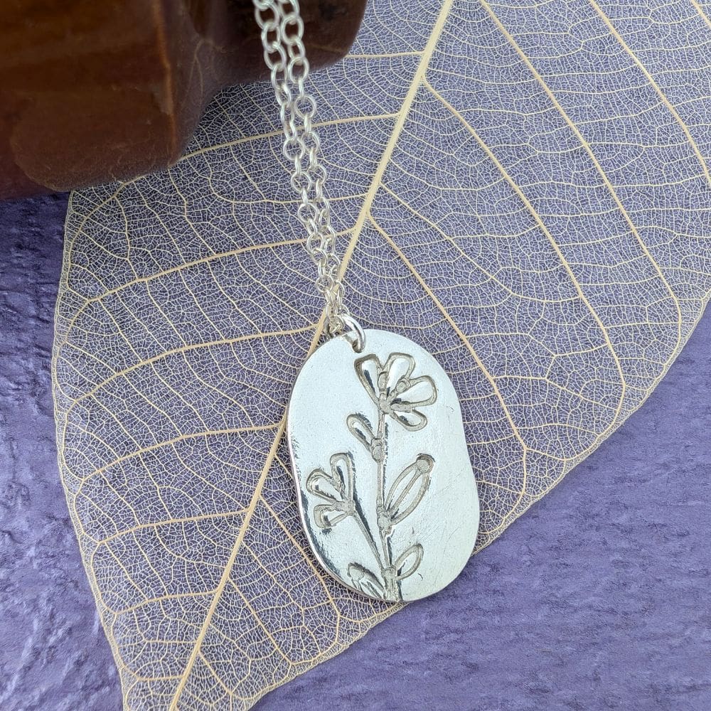 A fine silver dog tag shaped pendant with a wild flower imprint. Laid on a skeleton leaf with deep lilac background