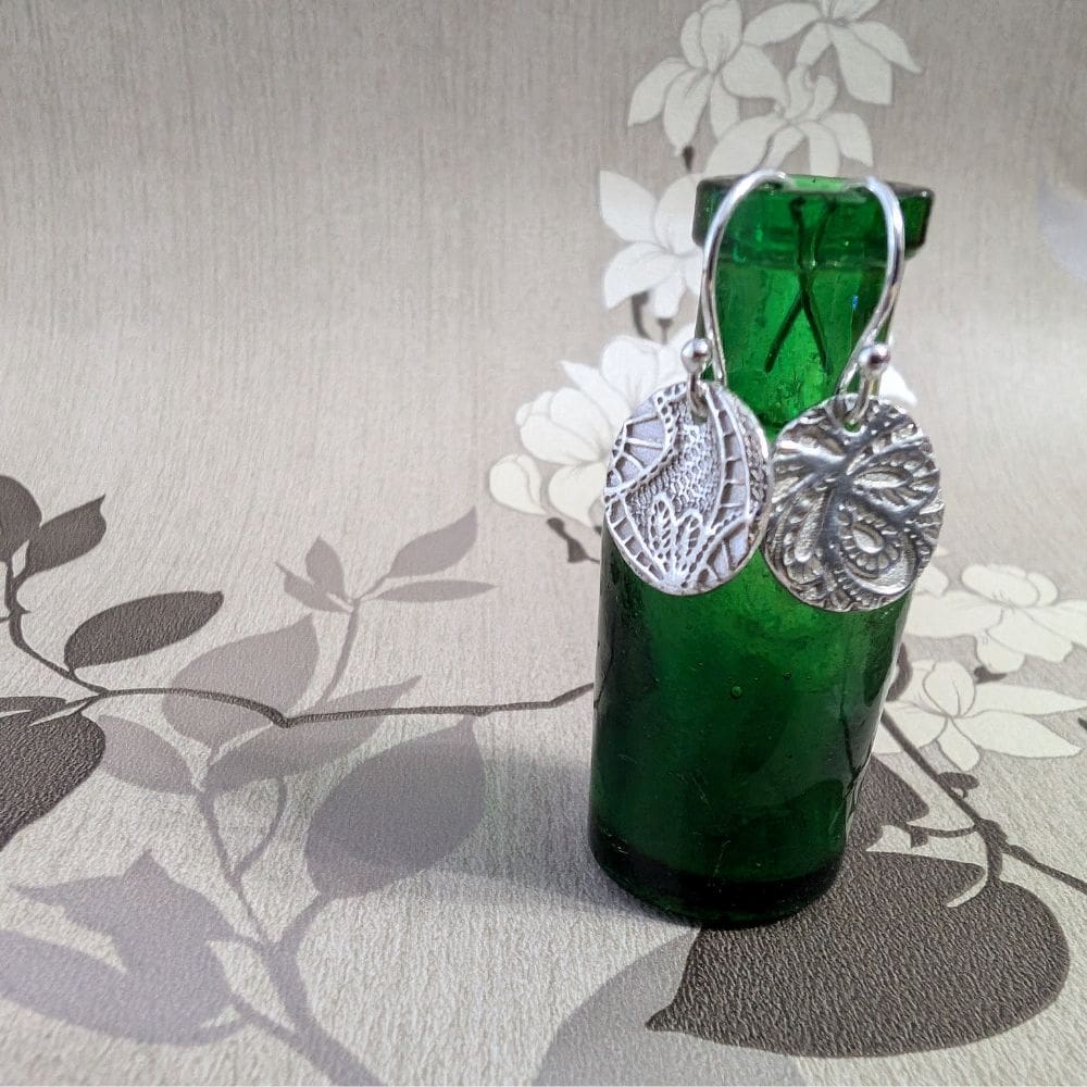 Silver drop earring with a lace texture are hung on a small vintage green bottle. The background is beige with pictures of oriental trees.