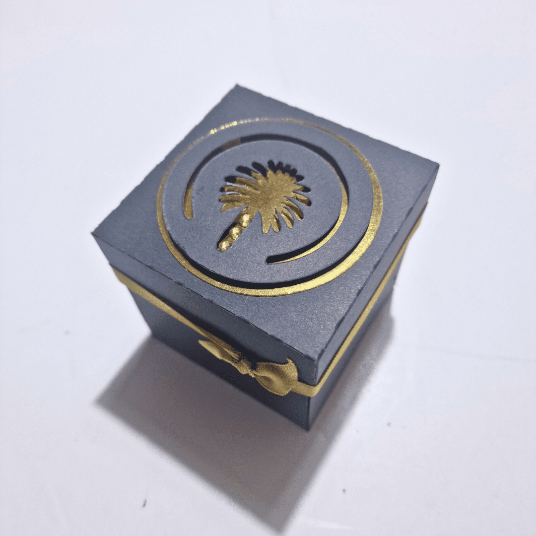 The image shows a small, square, dark-colored box with a golden design on the top and a golden ribbon tied around it. The design on the top features a palm tree within a circular pattern. The box appears to be made of paper or cardboard and has an elegant, luxurious appearance.