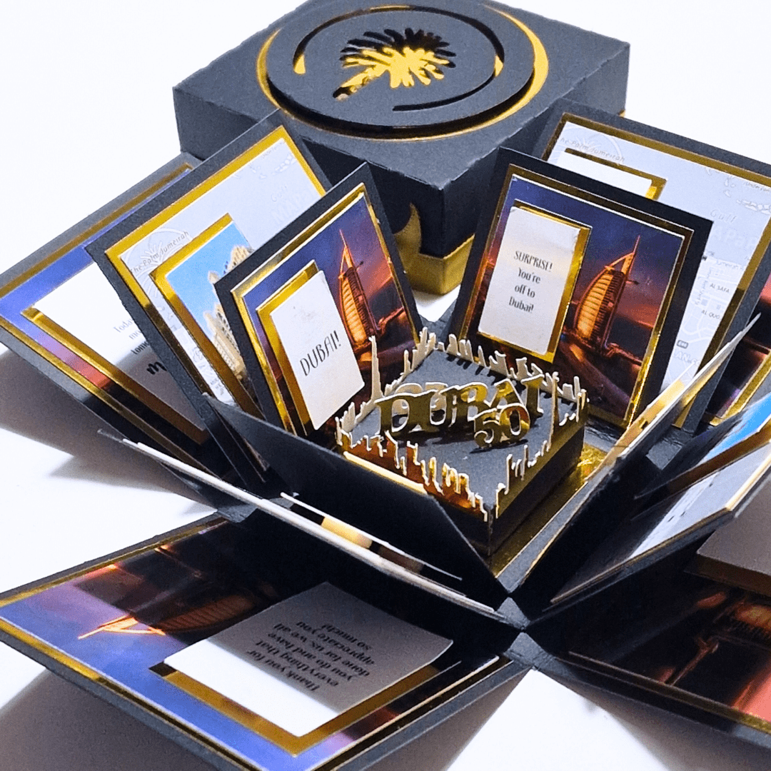 The image shows an intricately designed pop-up card or box that’s opened to reveal layers and compartments. The central piece is a 3D pop-up with the text "Dubai 50," surrounded by small candles or decorative elements. The panels display images of the Burj Al Arab hotel and messages like "SURPRISE!! You're off to Dubai!!" and "DUBAI!!" The box is black with gold accents, and the lid features a palm tree design.