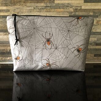 A large-sized makeup bag in grey featuring Halloween spiders, complemented by a black zipper and orange lining. Large internal slip pocket.