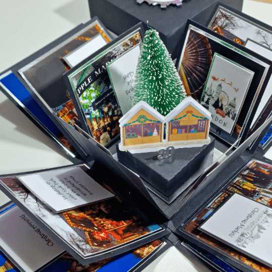 Black and Silver Christmas Markets Destination Trip Reveal Box. This fully opened view highlights all 12 panels, featuring a centerpiece with a Christmas tree and wooden-style chalets.