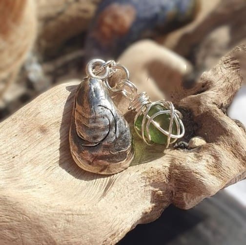 A silver mussel shell with a nugget of green sea glass wrapped in silver wire displayed on a piece of driftwood