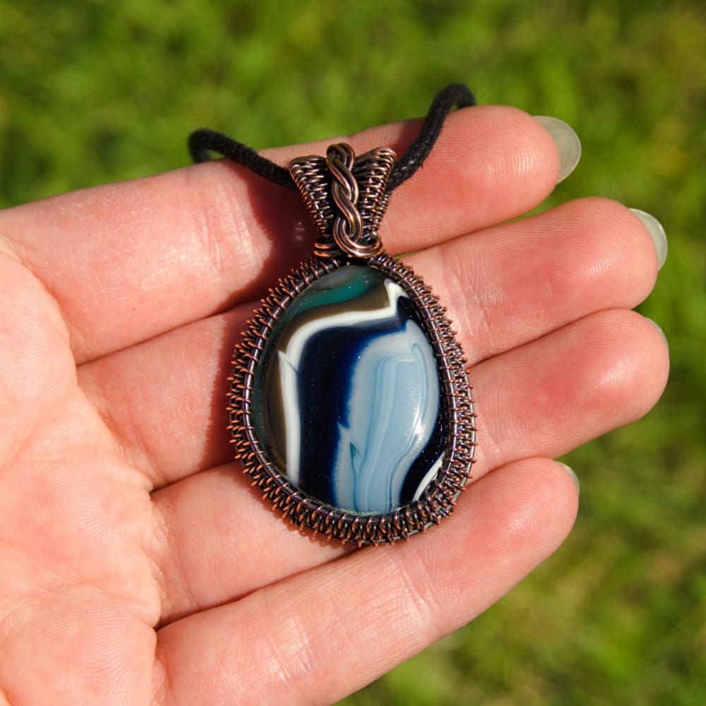 Hand holding a fused glass and copper pendant in blue, navy, white, brown and teal, by Oruki Design