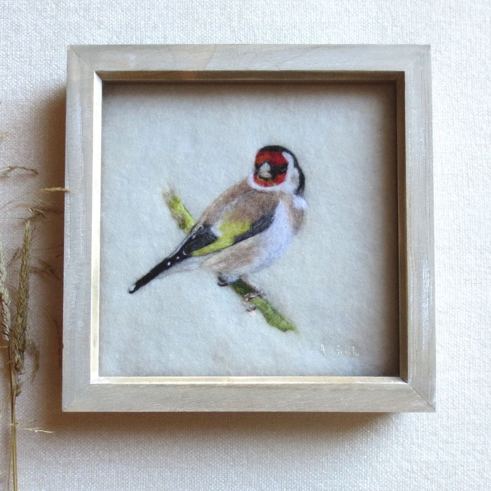 A small square picture of a Goldfinch bird made with wool. The Goldfinch is needle felted onto and cream wool background and is sat on a branch looking back over it's right wing. The picture is in a pale natural wood box frame.