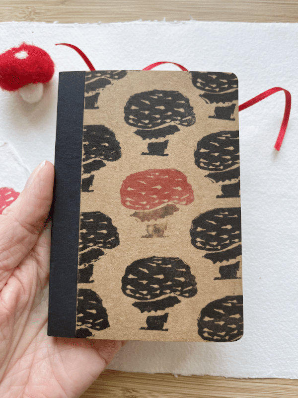 a mini mushroom notebook. It has a block printed design of a mushroom and is being held in a hand for scale