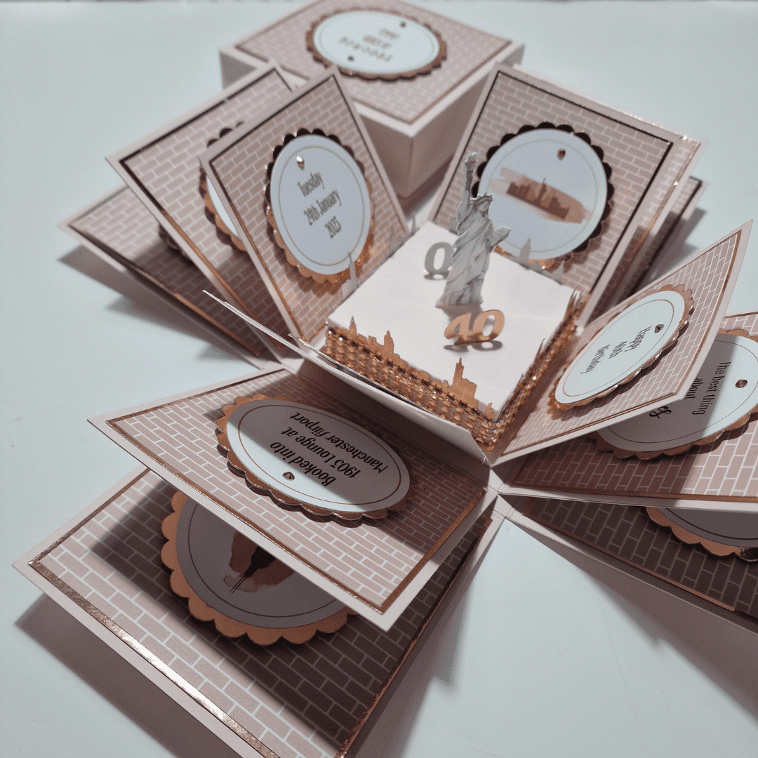 The image shows an intricately designed pop-up card or box that opens up to reveal multiple layers and elements. The central feature is a small 3D model of the Statue of Liberty, with the number "40" in front of it. Surrounding the central model are several panels, each decorated with a brick pattern and containing circular cutouts with text. The text on the panels includes dates and names, suggesting that this might be a commemorative or celebratory card, possibly for a 40th anniversary, Christmas gift or birthday.