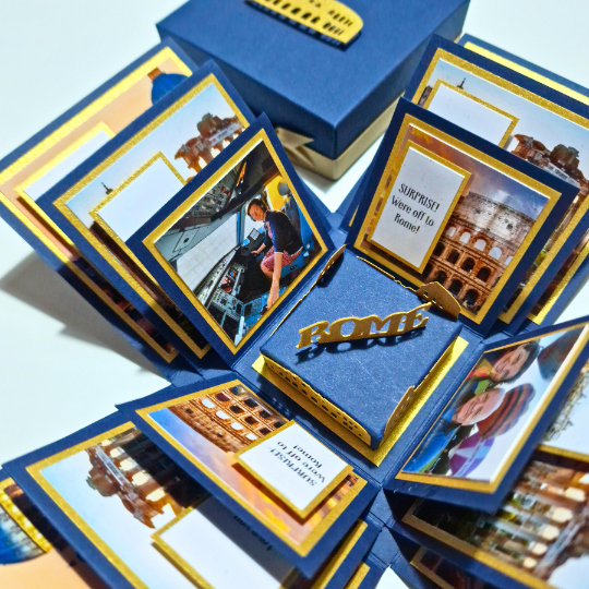 The image shows a pop-up card with multiple layers and sections. It's mainly blue and yellow, featuring photographs of famous Rome landmarks and a central 3D model of the Colosseum. One section reads, "SURPRISE! We're off to Rome!" There are also blurred faces of people in one of the photos. Quite the artistic and exciting way to announce a trip!