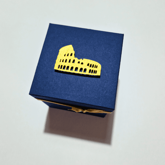 The image shows a small, dark blue box with a lid. On top of the lid is a golden cutout of the Colosseum, making it a visually appealing and thematic gift box, especially for someone interested in Roman history or architecture