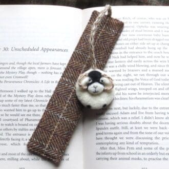 A brown checked wool tweed bookmark with a needle felted wool Swaledale sheep attached to the top with a jute string cord. The bookmark is resting on an open book.