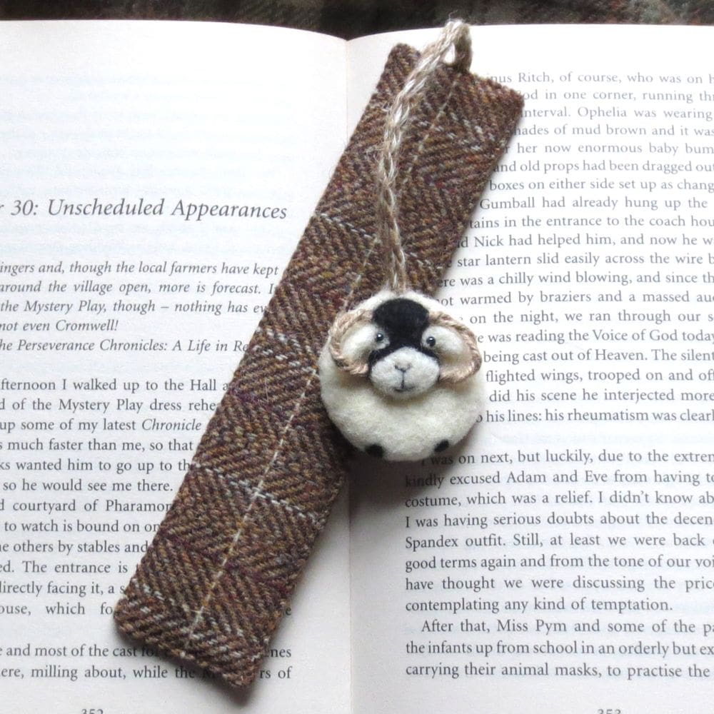 A brown checked wool tweed bookmark with a needle felted wool Swaledale sheep attached to the top with a jute string cord. The bookmark is resting on an open book.