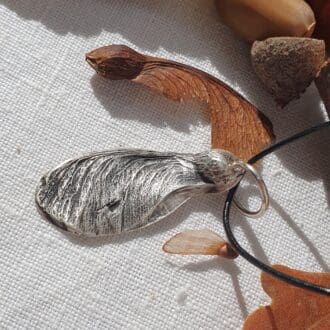 A silver sycamore seed with a leather cord surrounded by real sycamore seeds and autumn leaves on an off-white linen cloth