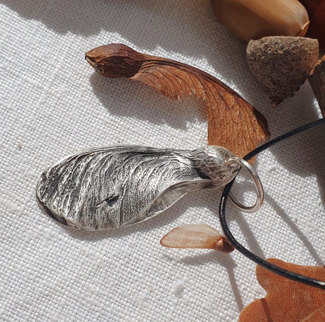 A silver sycamore seed with a leather cord surrounded by real sycamore seeds and autumn leaves on an off-white linen cloth