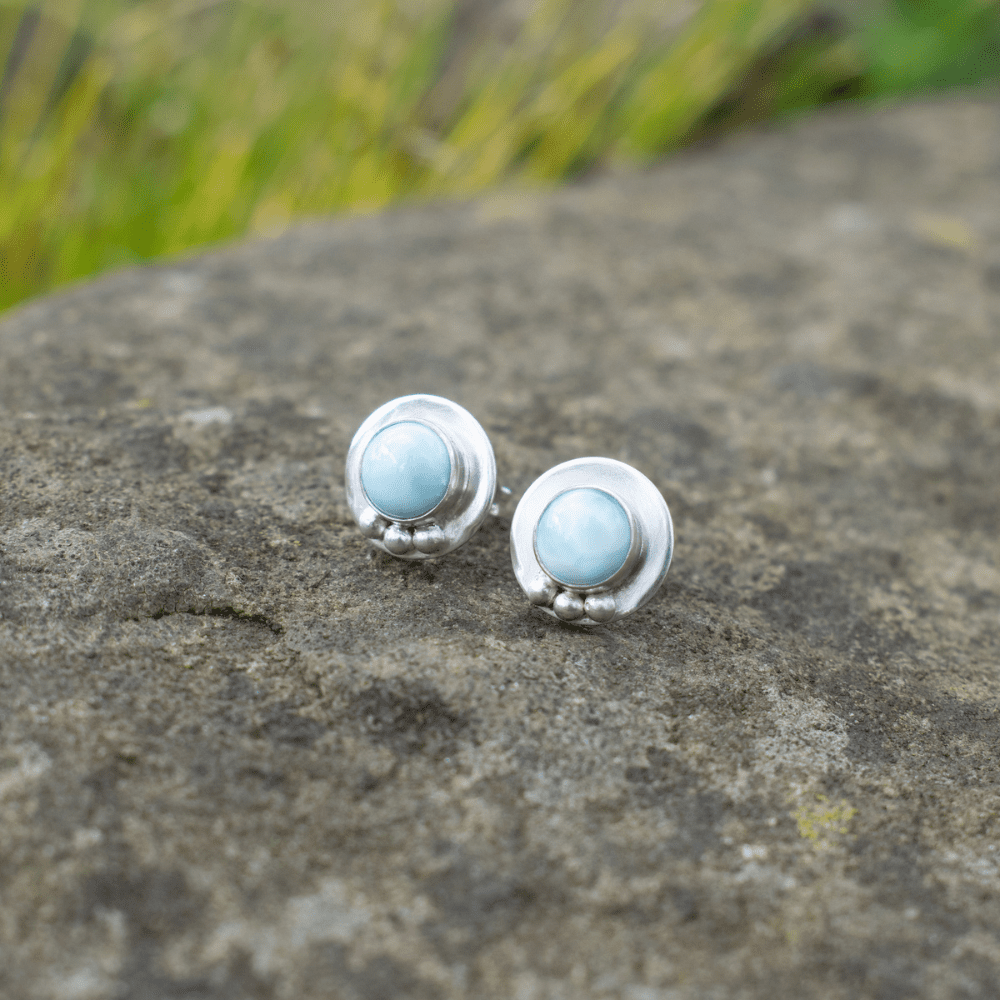 small round silver stud earrings with blue larimar stones set in the middle. Earrings rest on a large stone outside with plants in background