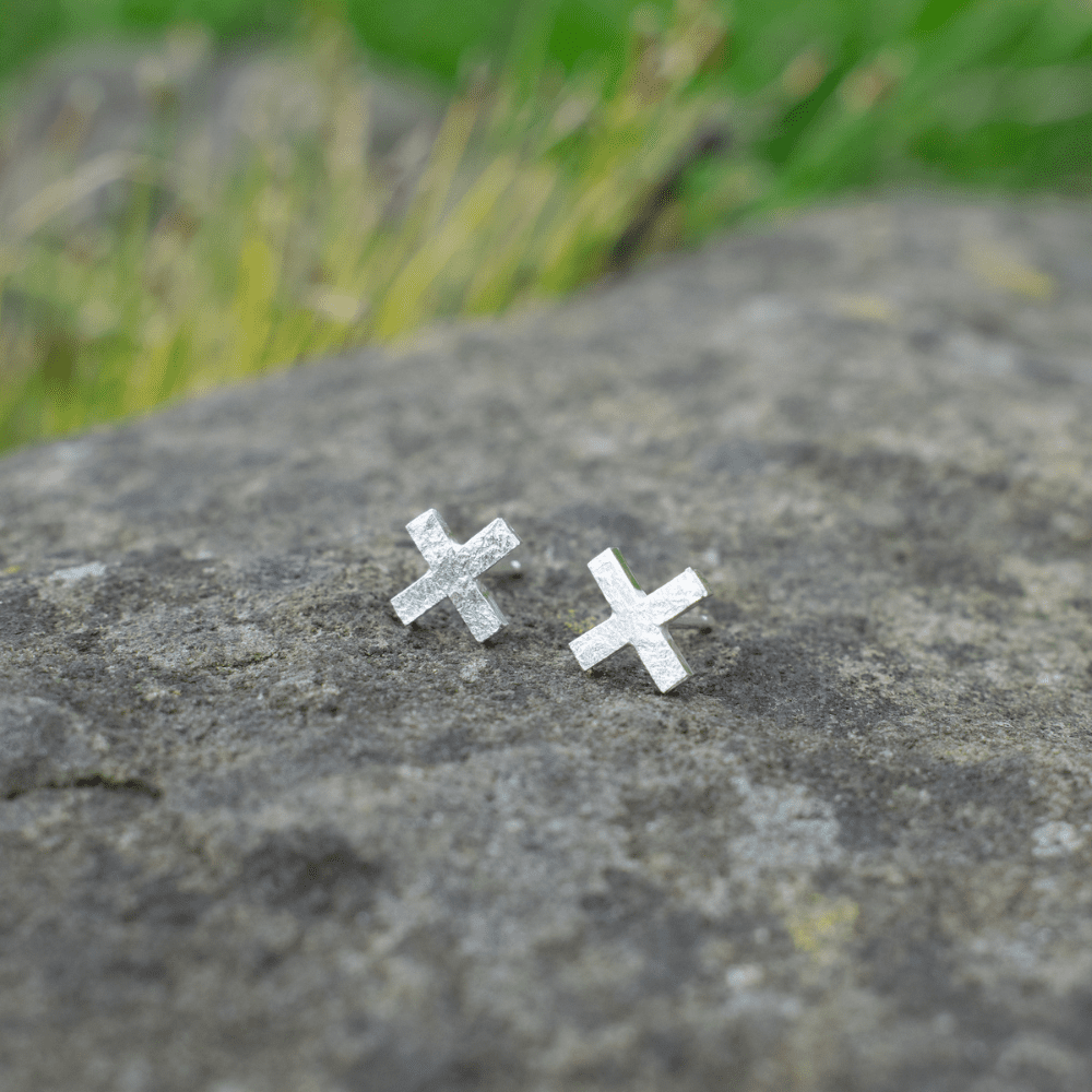 silver cross shaped stud earrings on stone with plant in background