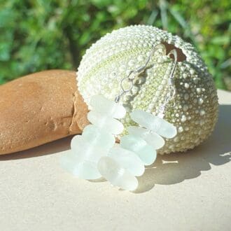 A pair of white sea glass earrings of various shades leaning against a sea urchin leaning against a pebble