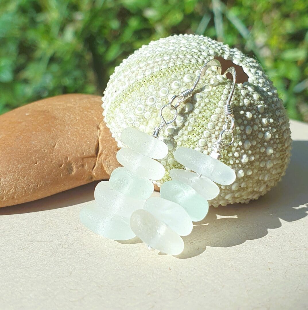 A pair of white sea glass earrings of various shades leaning against a sea urchin leaning against a pebble