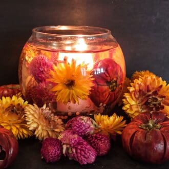 Autumnal themed botanical candle with rich coloured real flowers and pumpkins glows cosily against a dark background surrounded by dried botanicals.