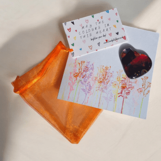 Handmade fused glass heart with multicoloured design, placed next to a handstamped card with a floral illustration and a quote saying "May the colours in this heart brighten your day." The set includes an orange organza bag, perfect for gifting.