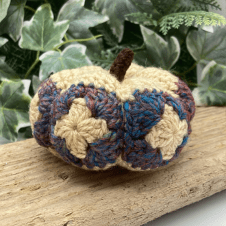 Its a picture of a crochet pumpkin made from granny squares. Its sitting on a piece of wood and behind it is a green leafy background