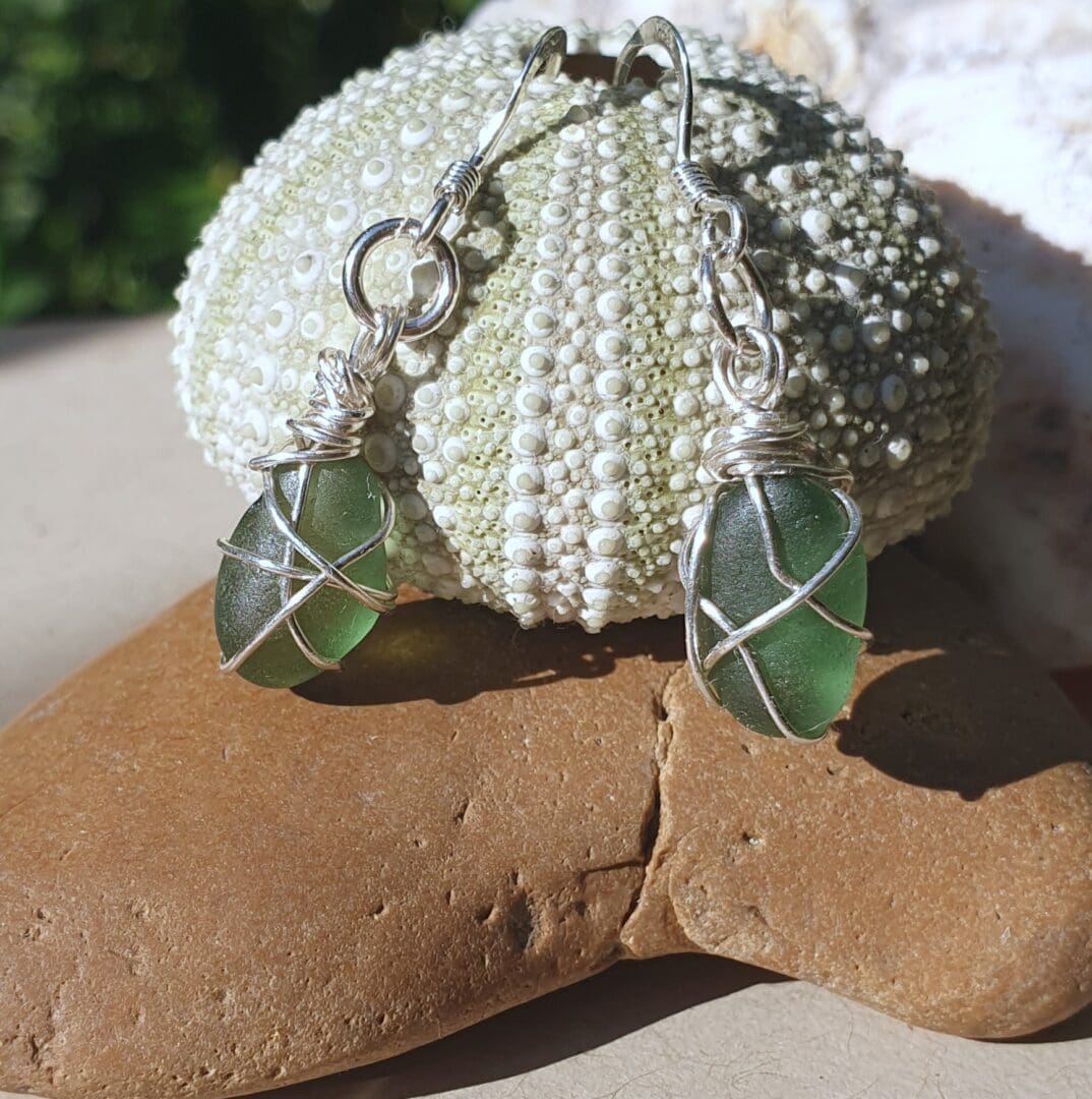 A small green sea urchin on a tan coloured stone, with green sea glass earrings wrapped in silver wire hanging from the urchin