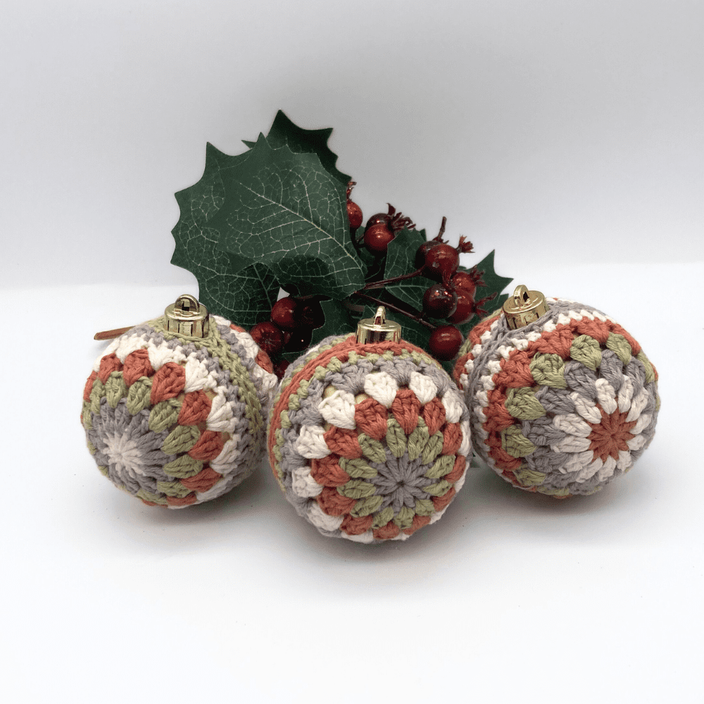 3 crochet covered baubles in earth tones. Behind them is a sprig of greenery with red berries on