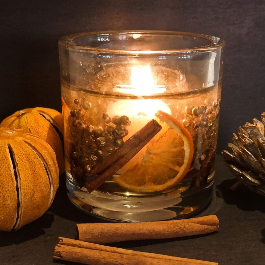 Orange spice festive candle glows warmly on a dark surface surrounded by cinnamon sticks, dried oranges and pine cones. The candle has gold berries, cinnamon sticks and orange slices encapsulated in a clear gel wax with a central scented soy wax candle.