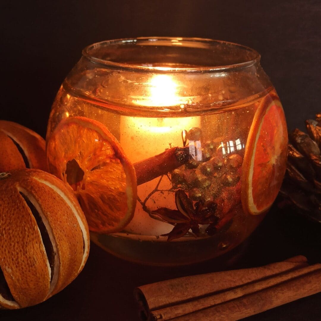 Orange spice scented botanical candle glows invitingly on a dark background surrounded by cinnamon sticks and dried oranges. The bowl shaped candle has inclusions of orange slices, cinnamon sticks and gold berries set into a clear gel wax. In the centre there is a scented white soy votive candle.