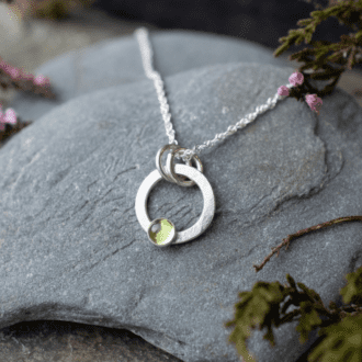 silver open circle pendant with green stone set at bottom. Necklace rests on a slate pebble with heather foliage around edge