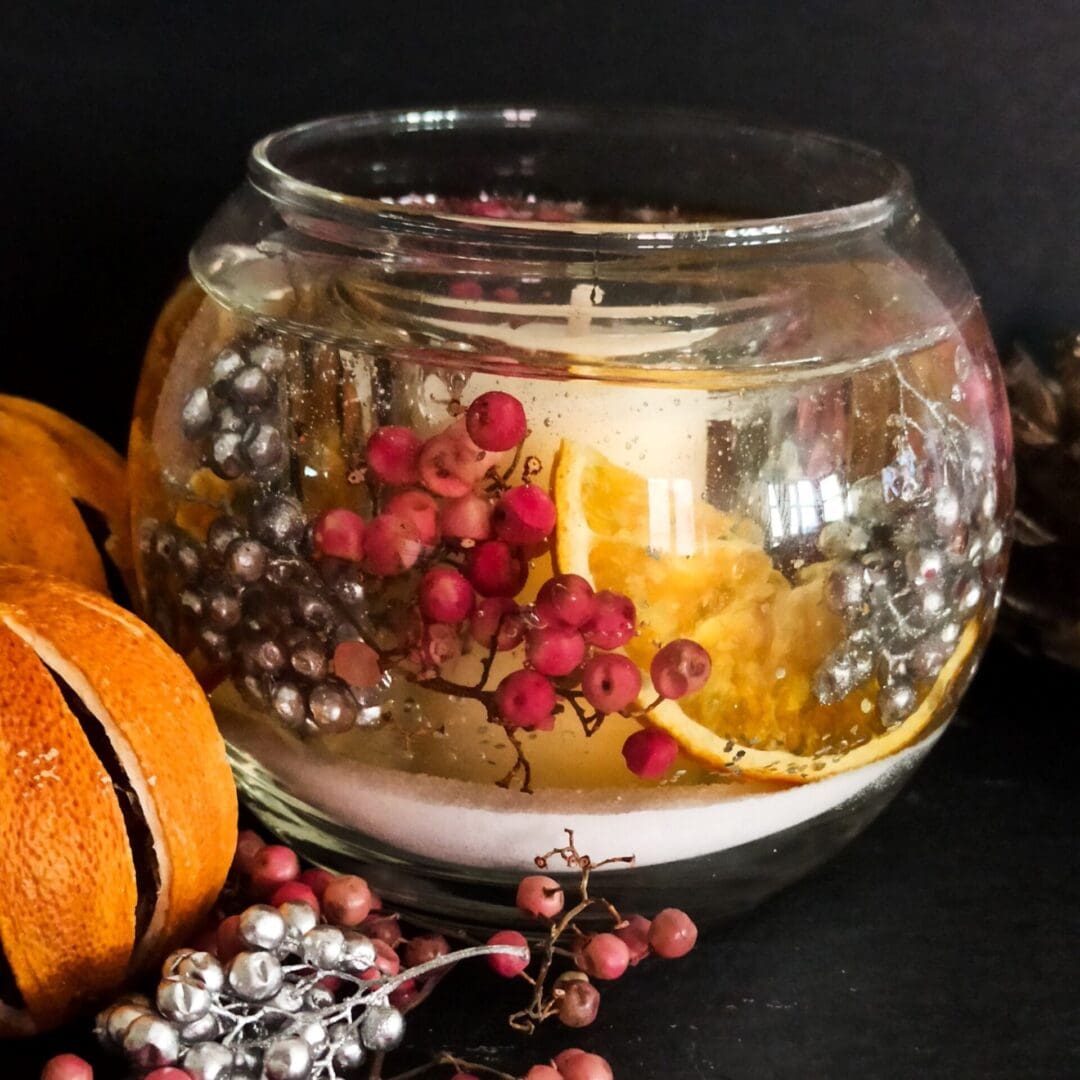 Sparkling cranberry luxury botanical candle rests on a dark background surrounded by pink and silver berries with whole dried oranges. The bowl shaped candle is decorated with cool snow like white sand and silver berries. Vibrant pink berries and orange slices provide warm colours. The botanicals are suspended in gel wax and a soy wax votive is placed centrally.