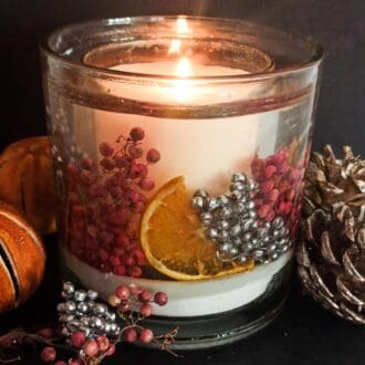 Sparkling cranberry scented luxury christmas botanical candle is shown on a dark background surrounded by berries and festive fruits. The candle contains a bed of white snow like sand on which are placed real silver and pink berries with vibrant orange slices all suspended in a clear gel wax. The central soy wax candle glows invitingly.