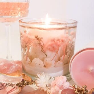 Pink champagne botanical statement candle exudes luxury and a hint of decadence glowing next to a glass of pink champagne and pink and white flowers.