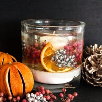 Sparkling cranberry christmas candle is placed on a dark background surrounded by pink and silver berries, dried oranges and frosted pinecones. The candle is filled with snow like soft white sand, metallic silver berries, pink berries and dried orange slices all suspended in a clear gel wax. A scented soy votive candle is placed centrally ready to be lit.