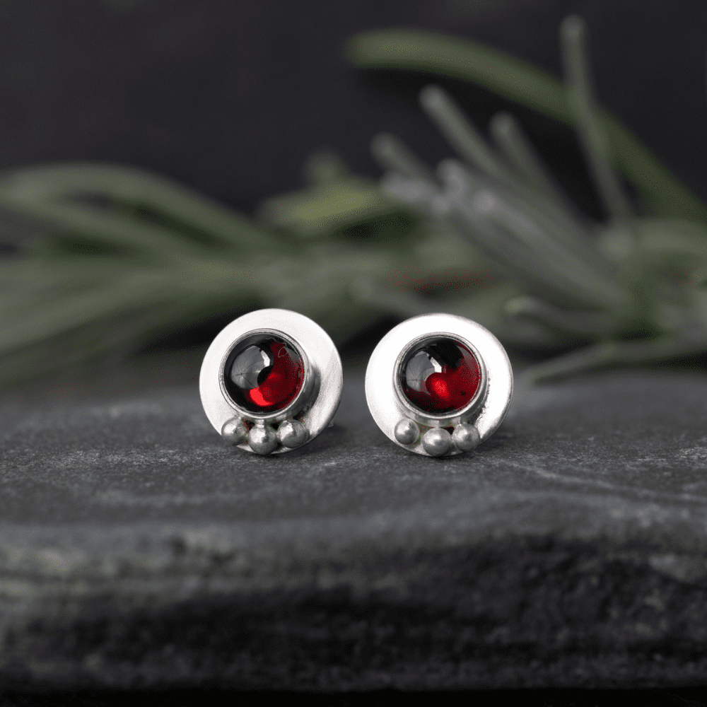 round silver stud earrings with red garnet stones in centre and three tiny silver pebbles at bottom. Earrings rest on a slate with foliage blurred in background.