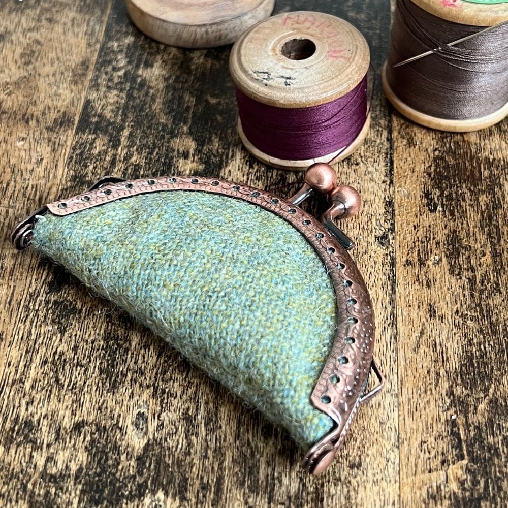 A small green purse clasp needle book sits on a wooden table surrounded by wooden vintage bobbins.