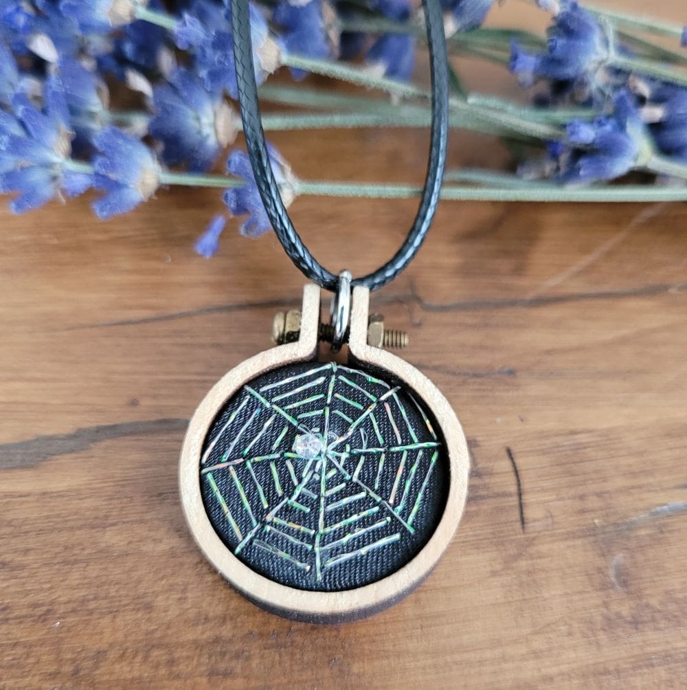 Hand embroidered mini hoop pendant of a spiders web with iridescent thread and a sparkly bead.