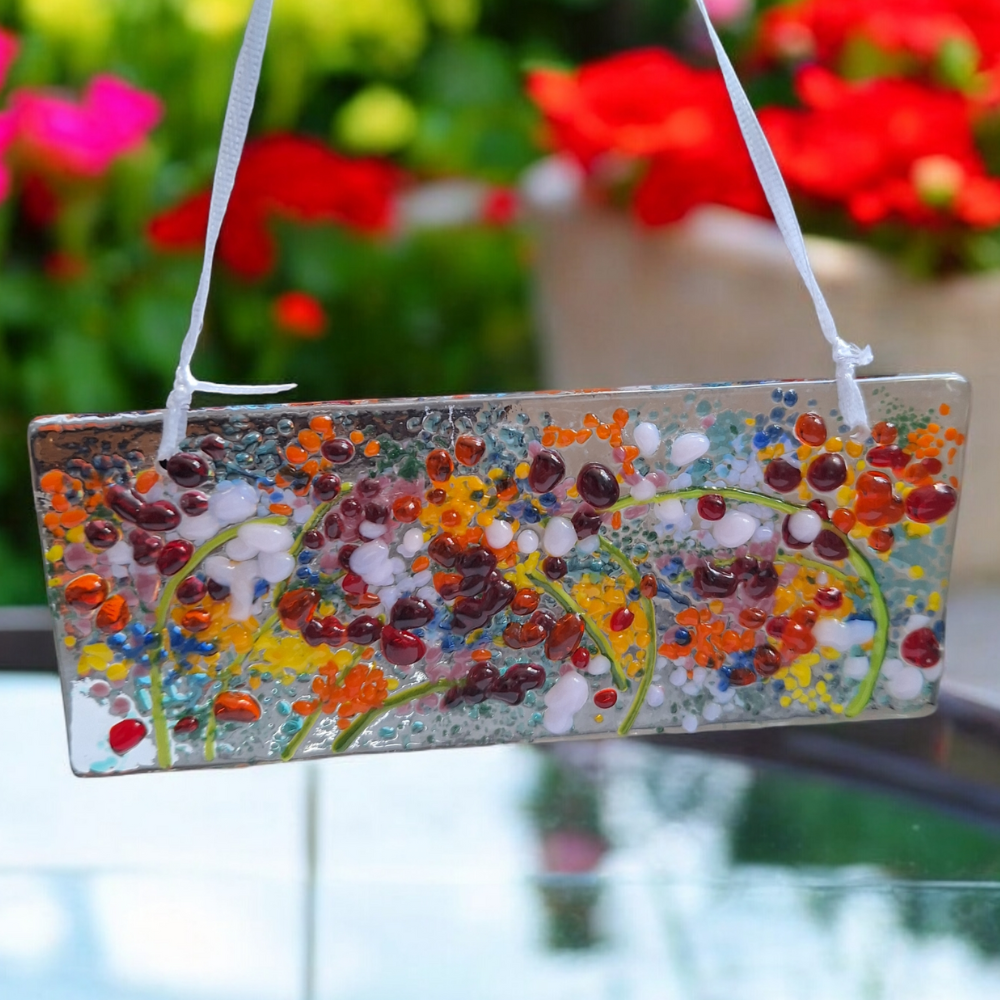 Handmade fused glass suncatcher featuring a colourful wildflower meadow design, hanging by a white ribbon. This unique glass ornament, measuring approximately 14 cm by 6 cm, showcases vibrant flowers in red, yellow, orange, and white, set against a textured clear glass background. Perfect for plant lovers, cottagecore decor, rustic home decor, or as a housewarming or anniversary gift.