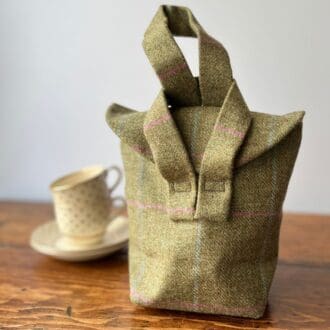 Picture shows a muted green wool check bag sitting on a wooden tabletop with a vintage style cup and saucer in the background. The bag is 1940s style and is fastened by linking one strap through the other.