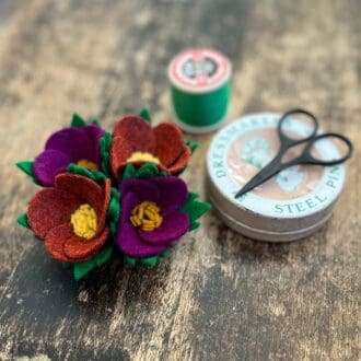 The picture shows a wool felt floral arrangement fashioned into a brooch sitting on a wooden table top. The brooch is made in the style of a 1940s 'make do and mend' design and features two purple and two bunt orange flowers. Next to the brooch is a reel of green thread used to hand stitch the leaves, a vintage tin of pins and a small pair of scissors.