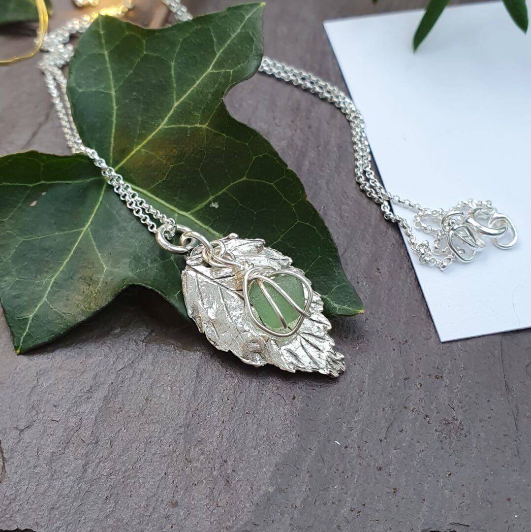 A silver rose leaf and wire wrapped green sea glass necklace displayed on an ivy leaf on slate