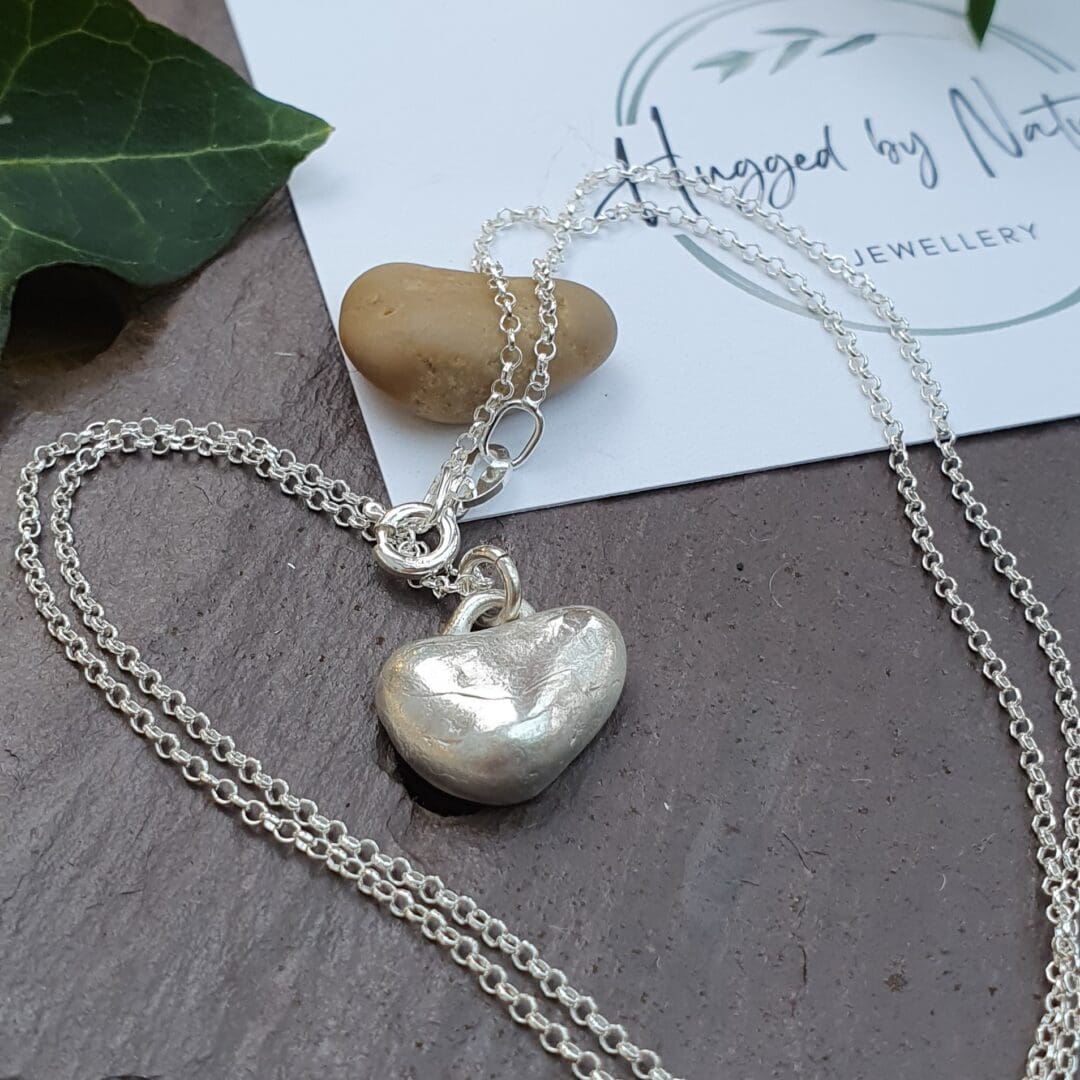 A small silver heart necklace next to the stone that inspired the heart displayed on a piece of slate.