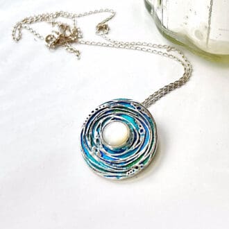 A round fine silver necklace that has been hand-painted with blues and turquoise. A mother of pearl stone is set in the centre and has a sterling silver chain.