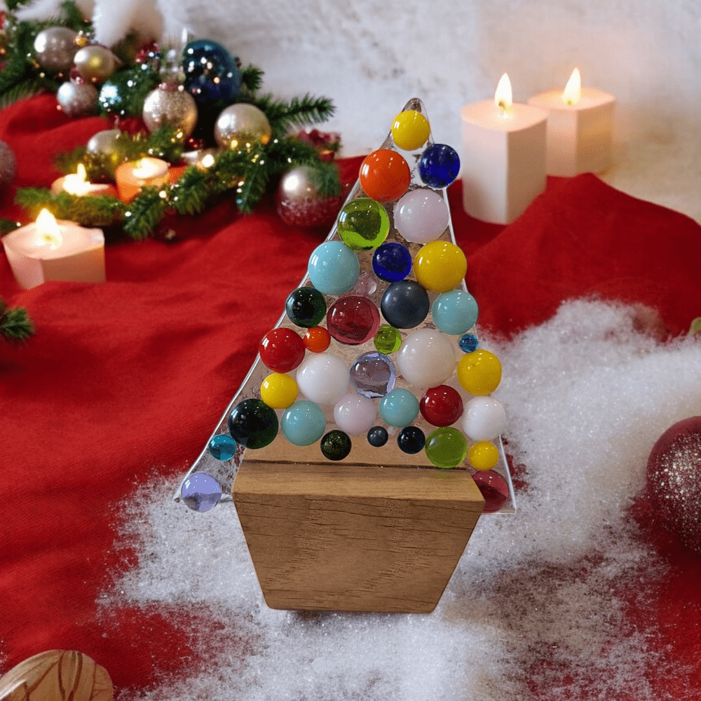 Multicoloured fused glass Christmas tree ornament with vibrant glass baubles on a wooden stand, set against a festive backdrop of candles, snow, and holiday decorations. Perfect handmade decoration for Christmas gifts, stocking fillers, Secret Santa, or Christmas Eve box ideas.