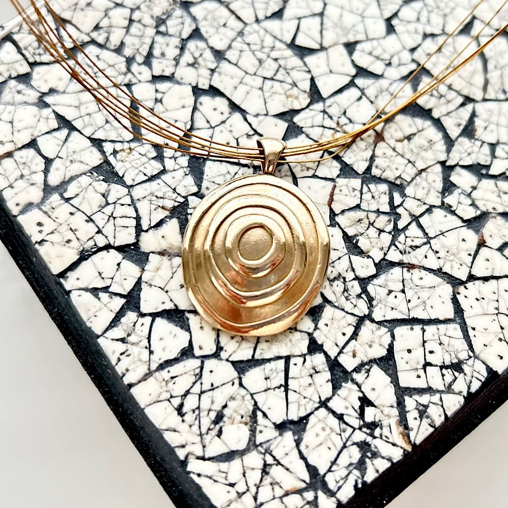 À bronze clay round pendant with a raised circle design, a gold coloured stainless steel stranded collar . Sat on a black-out and White pattern box.