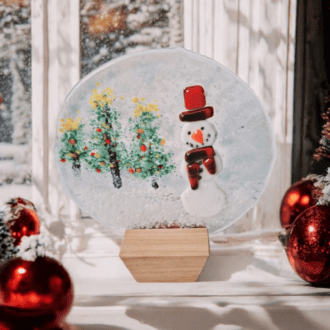 A festive fused glass snowman globe with a wooden stand. The globe features a snowman with a red hat and scarf, surrounded by a snowy scene with Christmas trees. Perfect for winter home decor and as a unique Christmas gift.